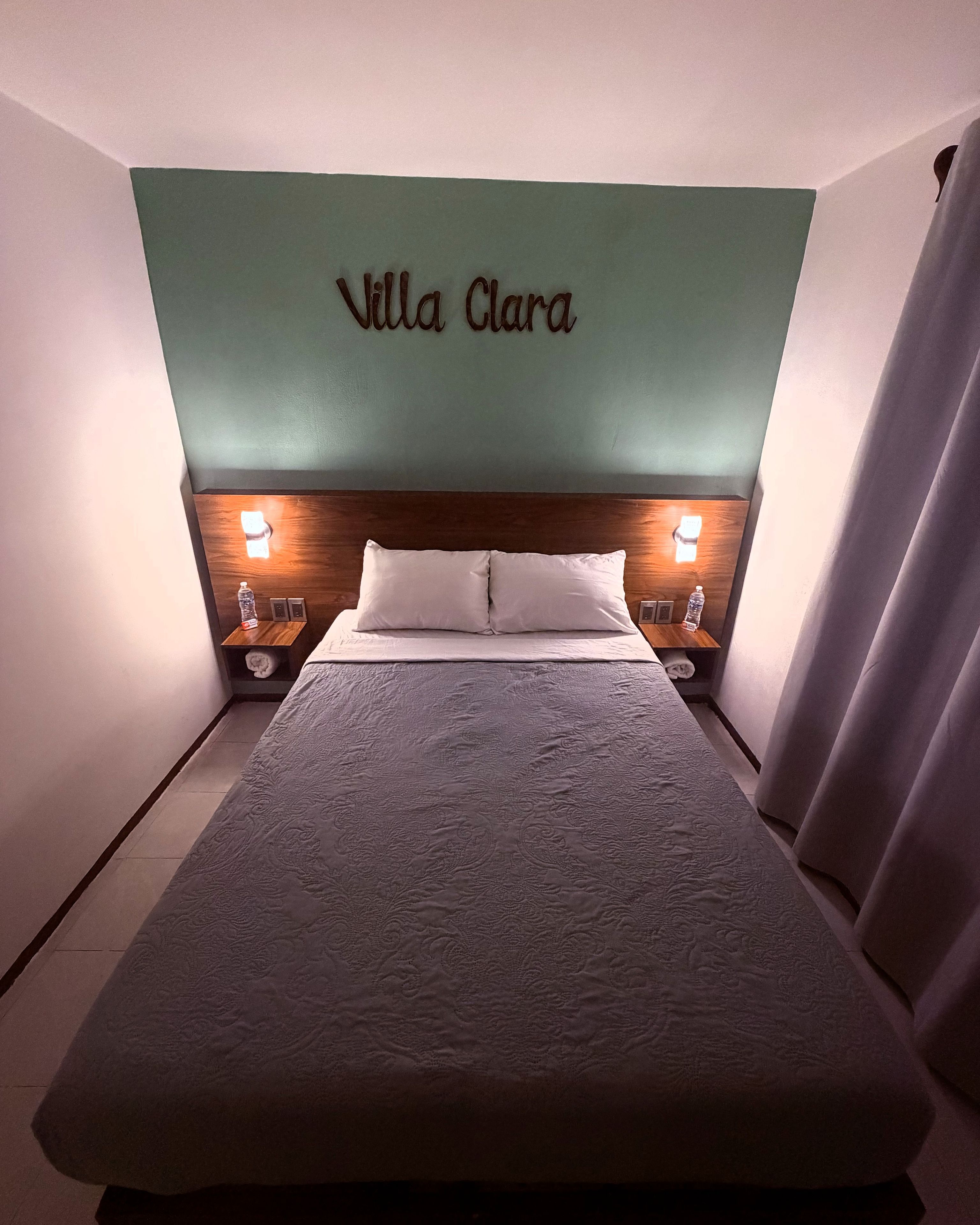 A hotel room with a bed, two bedside tables, a headboard with the name 'Villa Clara' on it, and a curtain on the right side.
