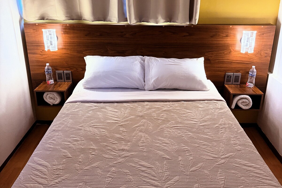 Habitación de hotel con una cama grande, dos mesitas de noche con lámparas, un cabecero de madera y vistas al exterior a través de cortinas.