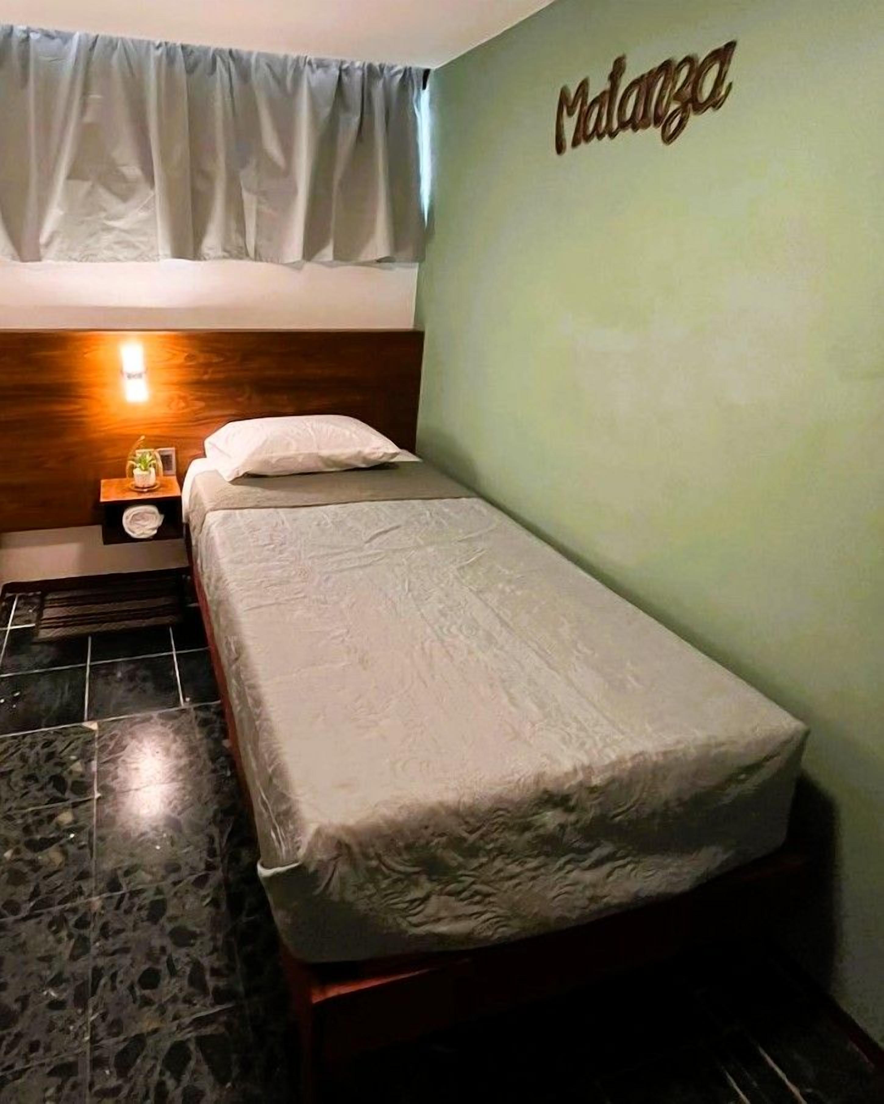 A single bed with a headboard, a bedside table with a lamp, and a wall with the name 'Malanga' in the corner.