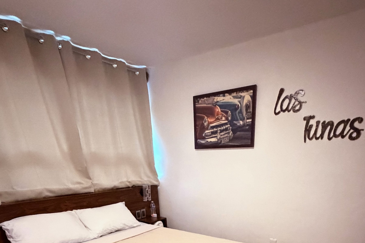 Una cama bien hecha en una habitación de hotel, con cabecera, mesita de noche, una botella y un cuadro con texto en la pared.
