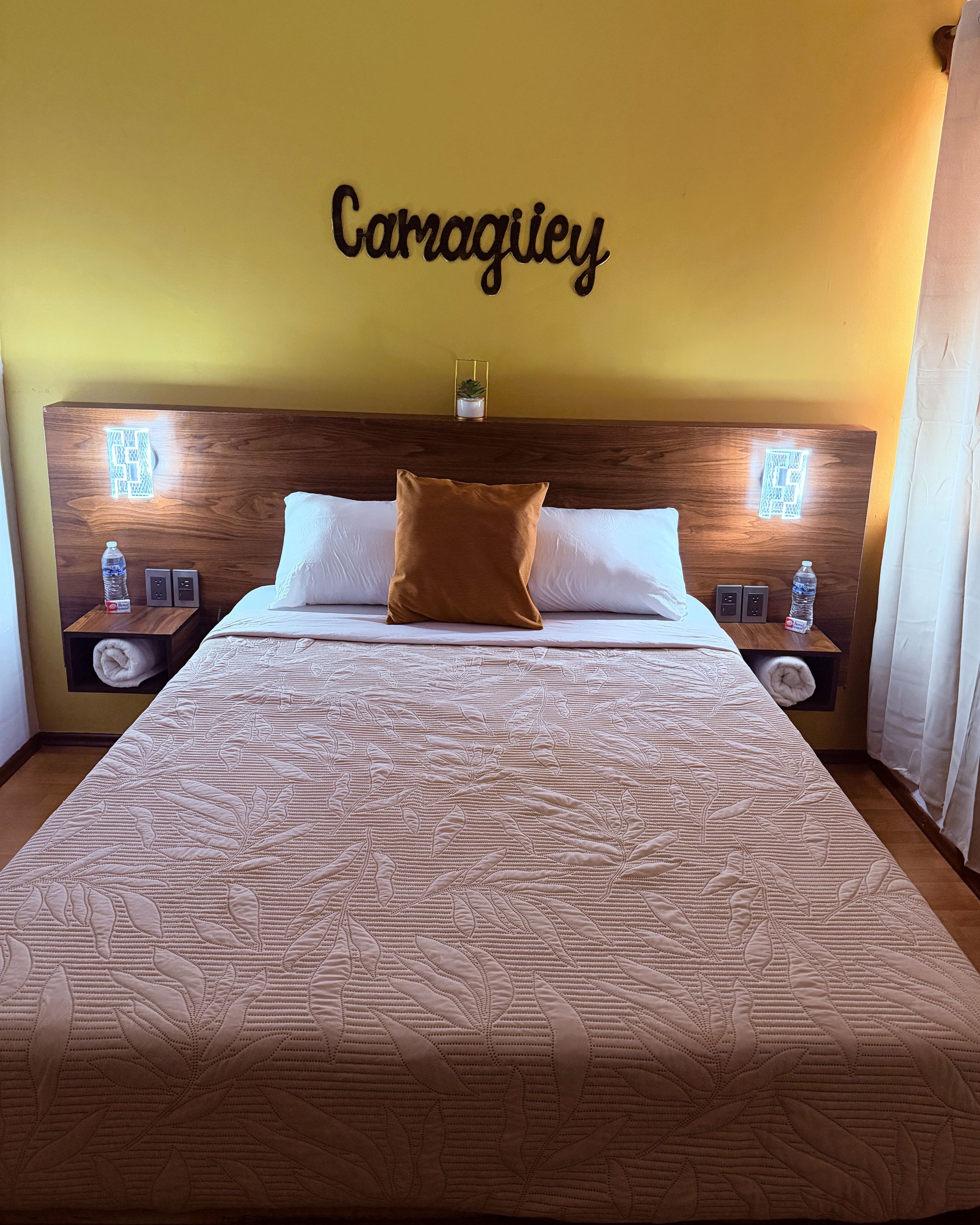 A hotel room with a large bed, two bedside tables, a headboard with a name plaque, and a small potted plant.