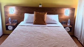 A hotel room with a large bed, two bedside tables, a headboard with a name plaque, and a small potted plant.