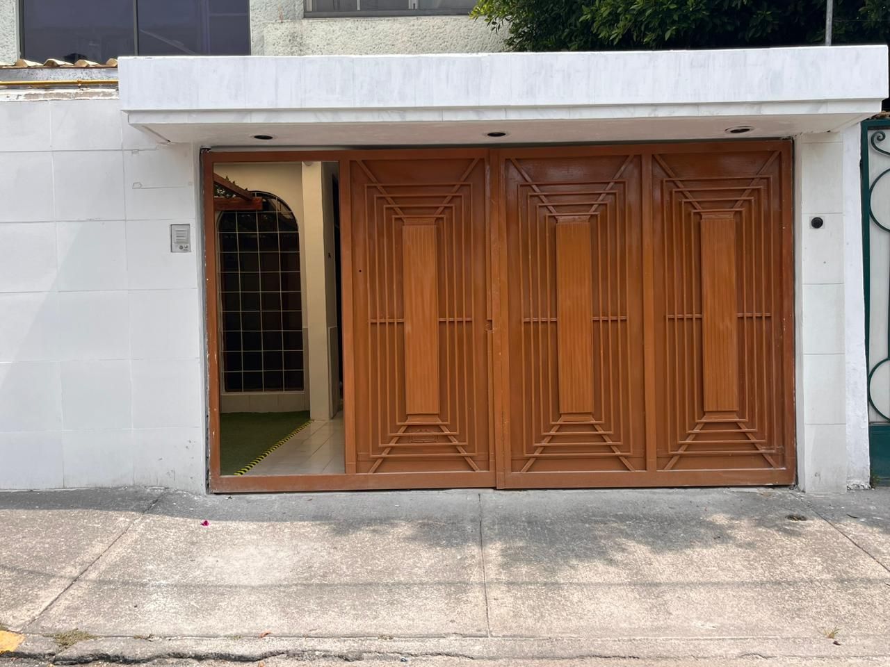 A double wooden door with a geometric pattern, set in a white wall with a metal gate to the right.