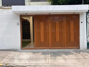 Una puerta doble de madera con un diseño geométrico, en una pared blanca con un portón metálico a la derecha.
