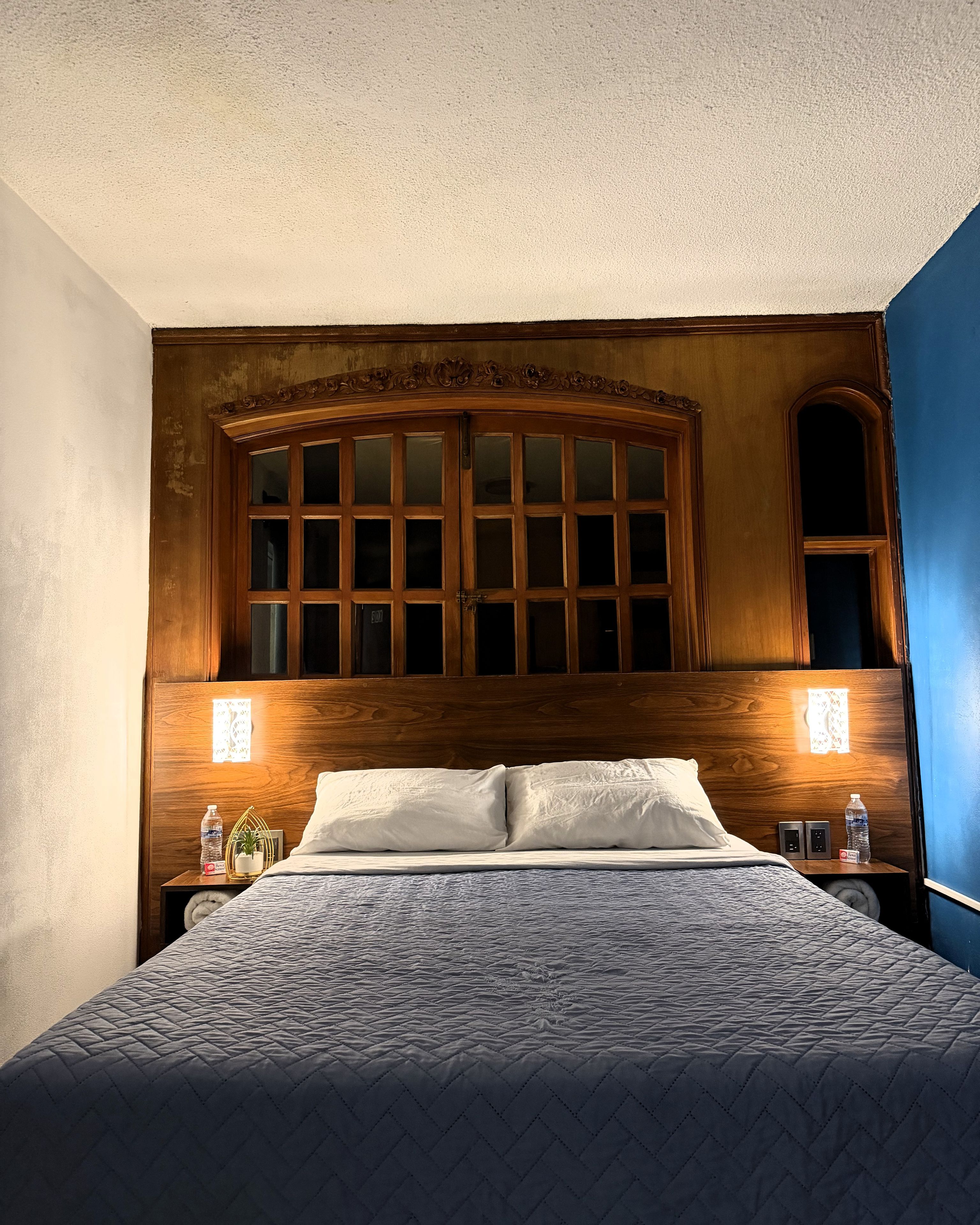 A hotel room with a large bed, wooden headboard, two bedside tables, and a nightstand with a lamp.