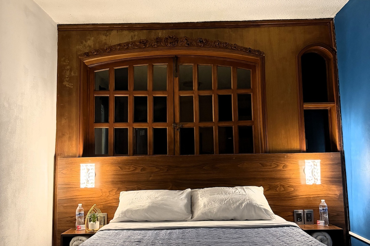 Una habitación de hotel con una cama grande, cabecera de madera, dos mesitas de noche y una mesita con lámpara.