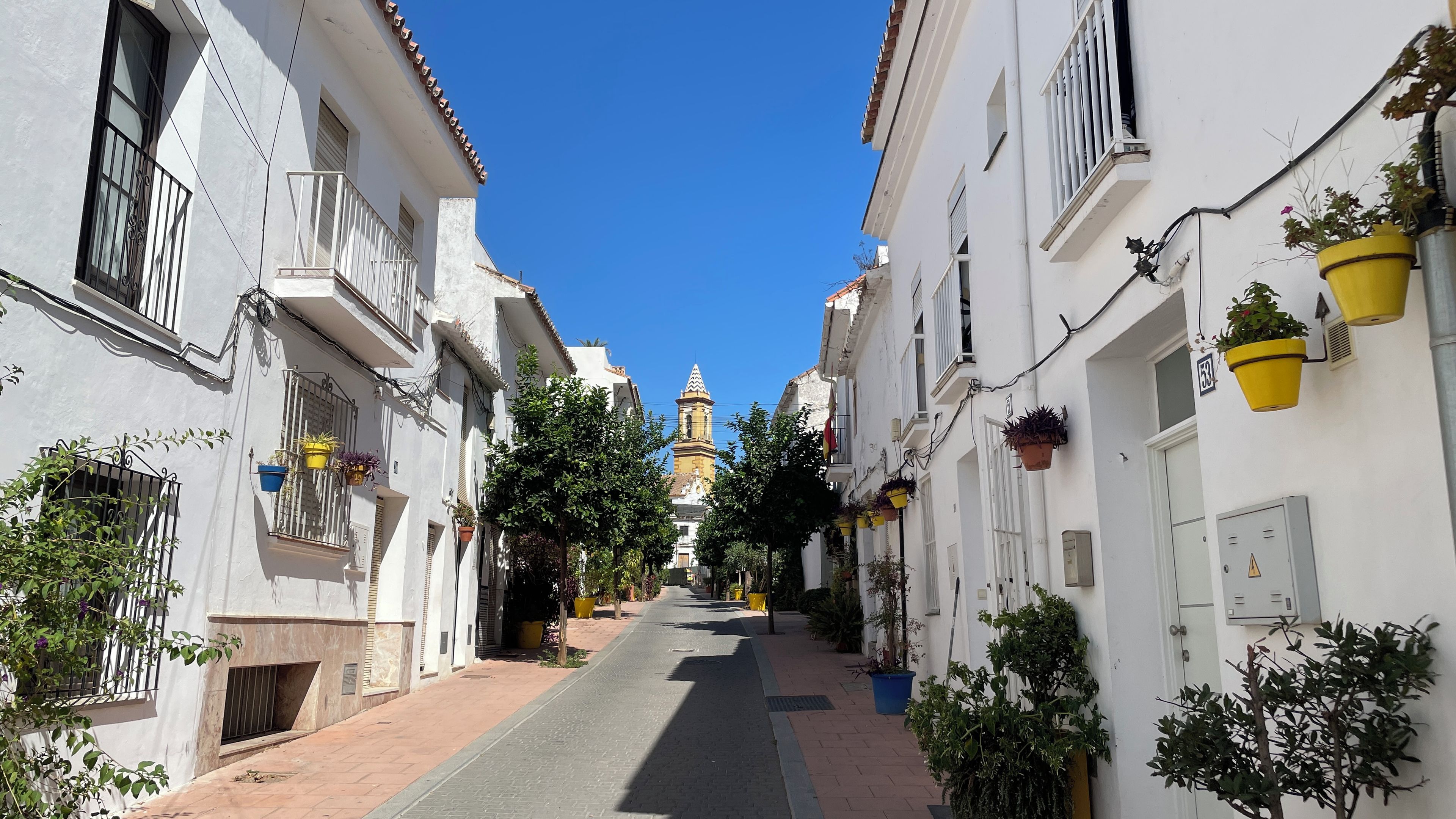 A narrow street lined with white buildings, some with balconies and potted plants. A tower with a clock is visible in the distance.