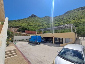 Ein Parkplatz mit einem blauen Lieferwagen und einem silbernen Auto, ein Gebäude mit einer überdachten Einfahrt und einem Bergpanorama im Hintergrund.