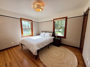 Un dormitorio con cama, mesitas de noche, un tapete y dos ventanas con marcos de madera.