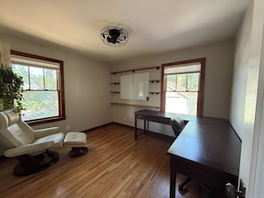 A room with a wooden floor, a desk, a chair, a ceiling fan, and a window with a view of greenery.