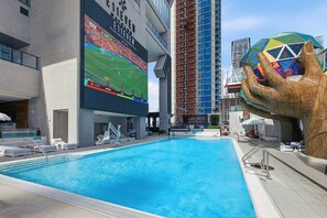 A swimming pool with lounge chairs, a large digital screen displaying a sports event, and a unique sculpture. - E11EVEN Skyline Studio | 33rd Floor City Views (Miami)