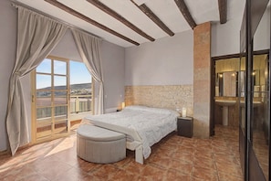 A bedroom with a stone headboard, a large window with curtains, a bedside table, and a round ottoman. - Vacation Apartment 'Apartamento Buen Lugar' with Sea View and Balcony (Buenlugar)