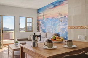 Zona de menjador amb taula de fusta, cafetera, un bol de fruita i un quadre amb una vista de la ciutat a la paret.