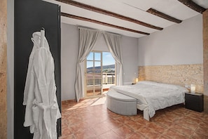 Un dormitori amb un llit, una finestra amb cortines, una paret de pedra i un paviment de rajoles.