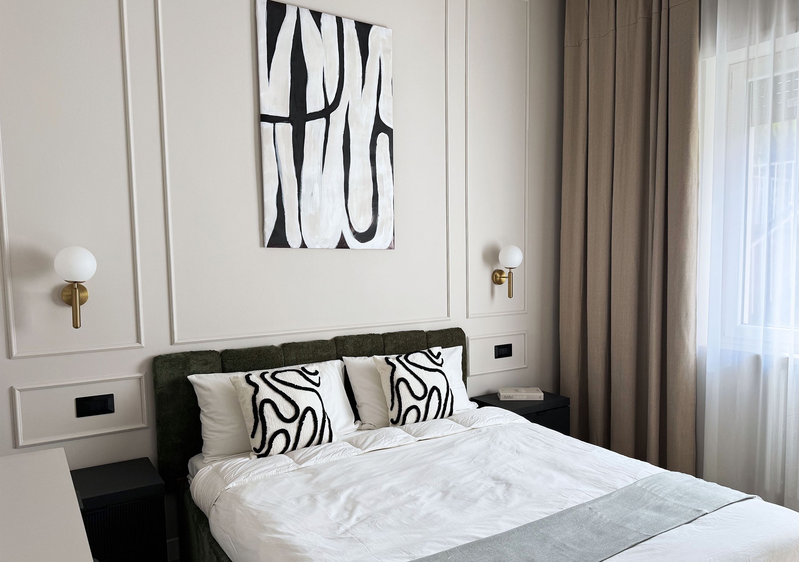 Habitación de hotel con cama, un cuadro abstracto grande, dos apliques de pared y una ventana con cortinas.