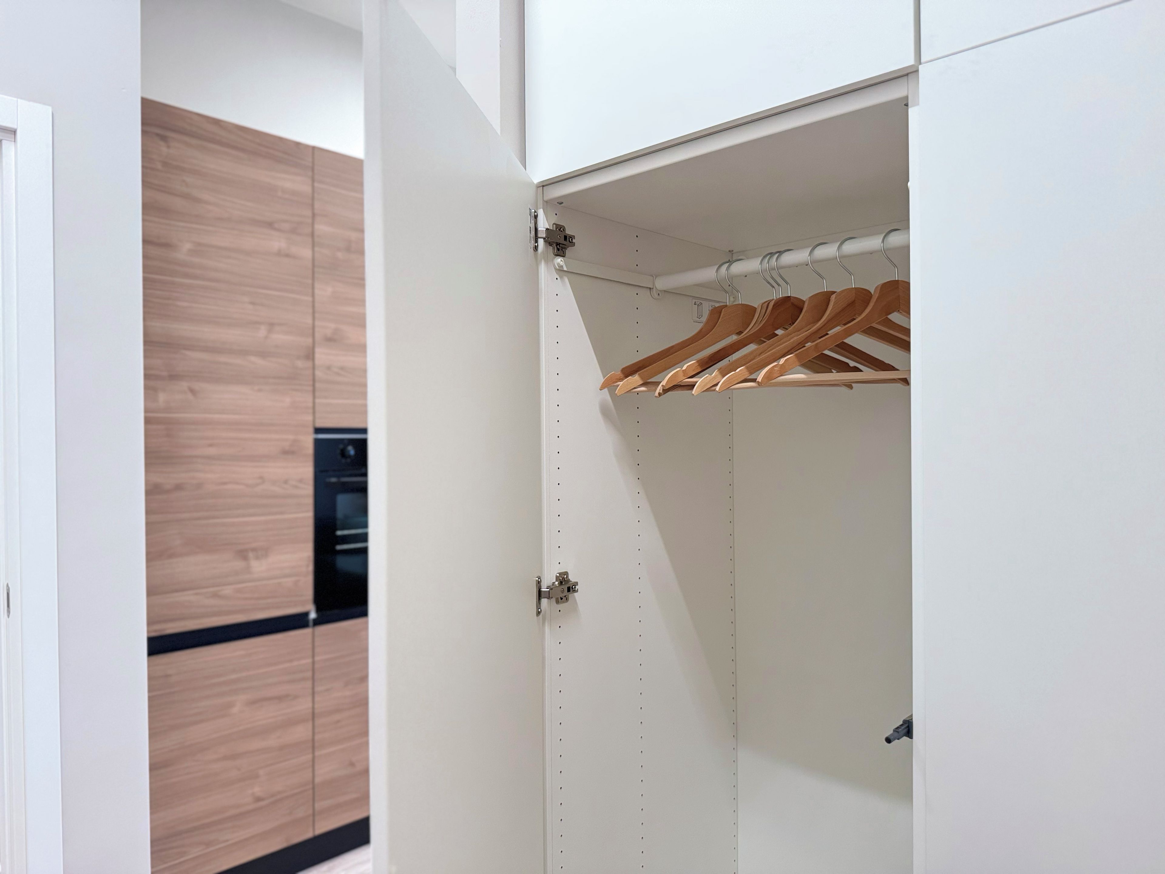 Un armario blanco con perchas de madera, un picaporte visible y una cocina al fondo con armarios de madera y encimera negra.