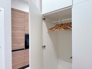 A white wardrobe with wooden hangers, a visible door handle, and a kitchen area with wooden cabinets and a black countertop in the background.