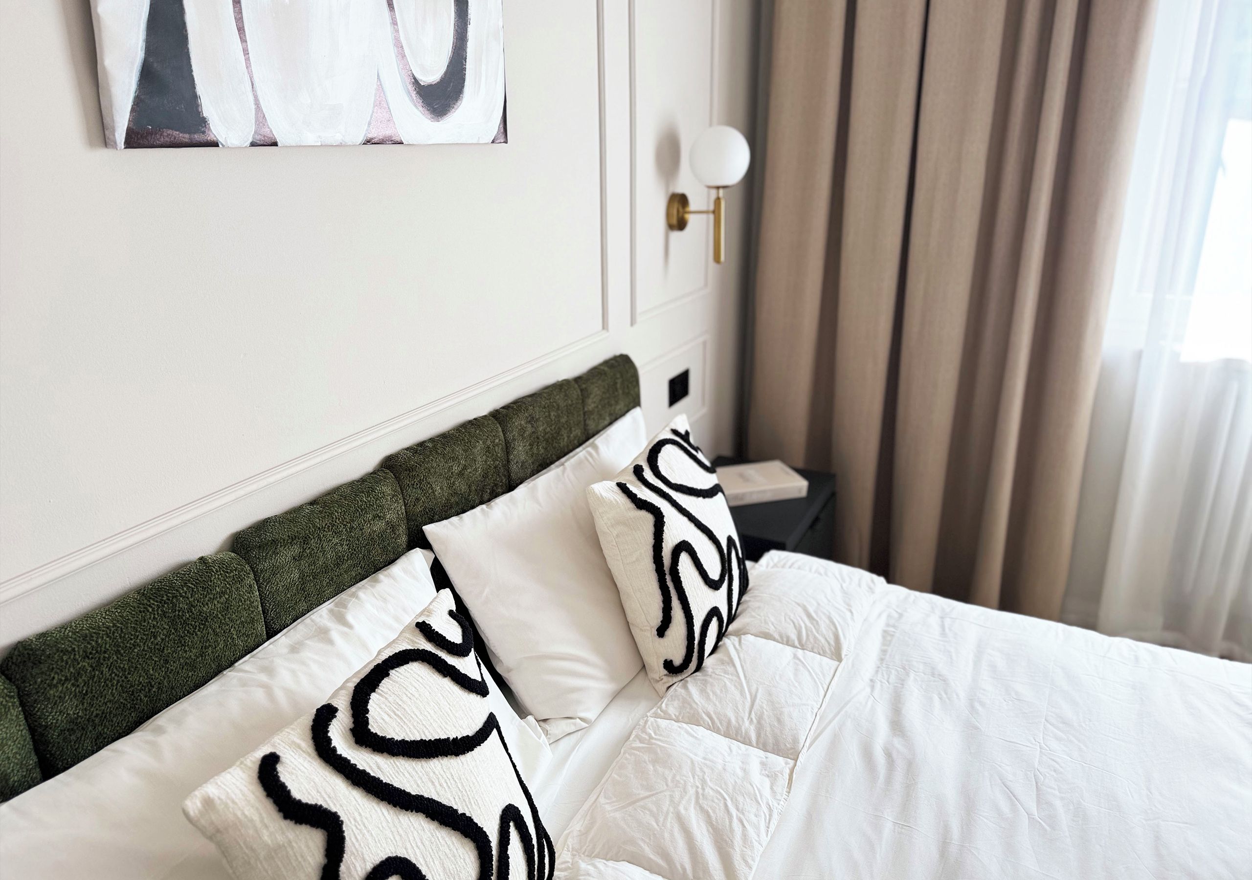 A neatly made bed with white bedding and a green headboard, a bedside table with a lamp, and a painting on the wall.