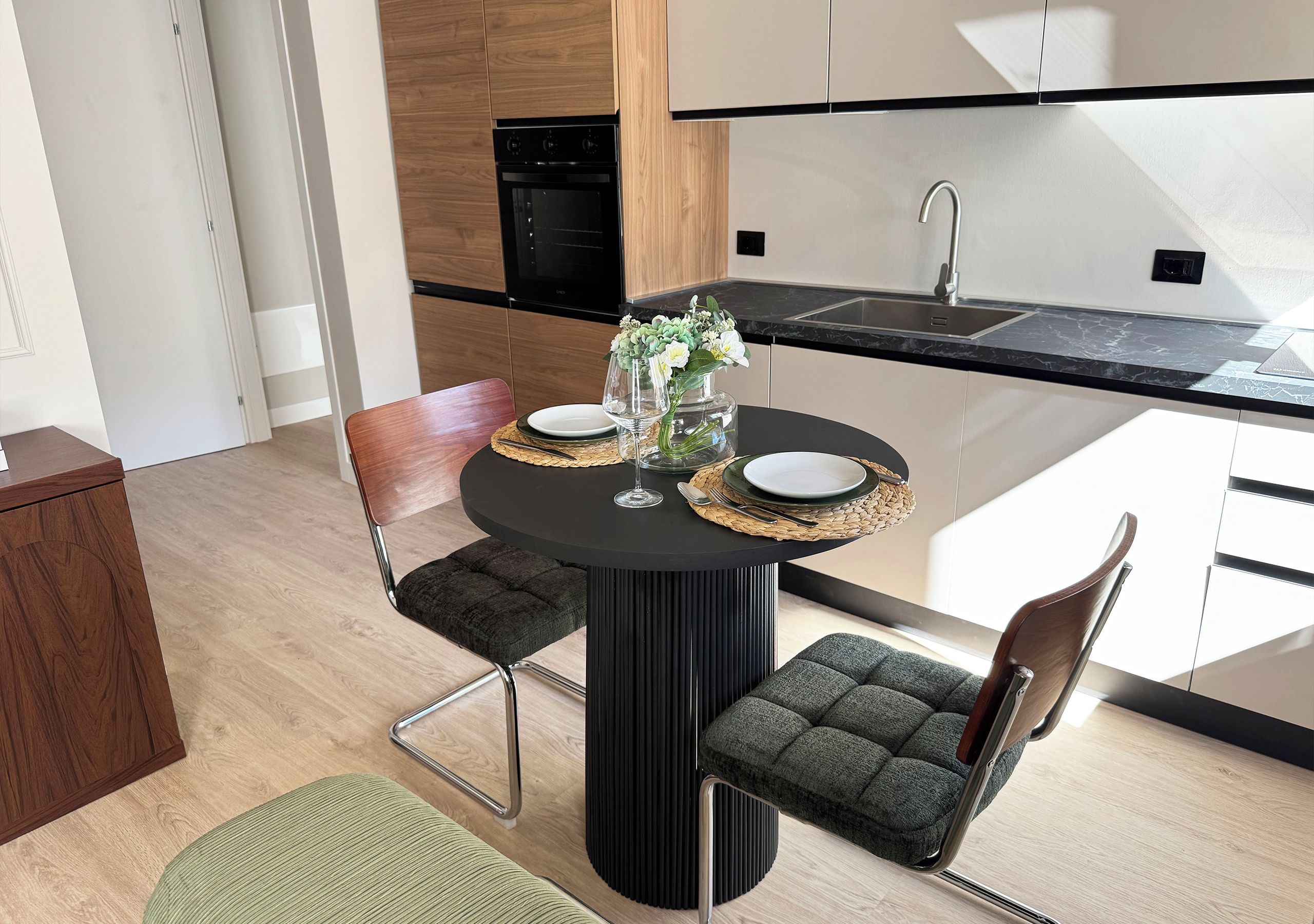A modern kitchen with a dining area, featuring a round table with chairs, a vase with flowers, and a black countertop.