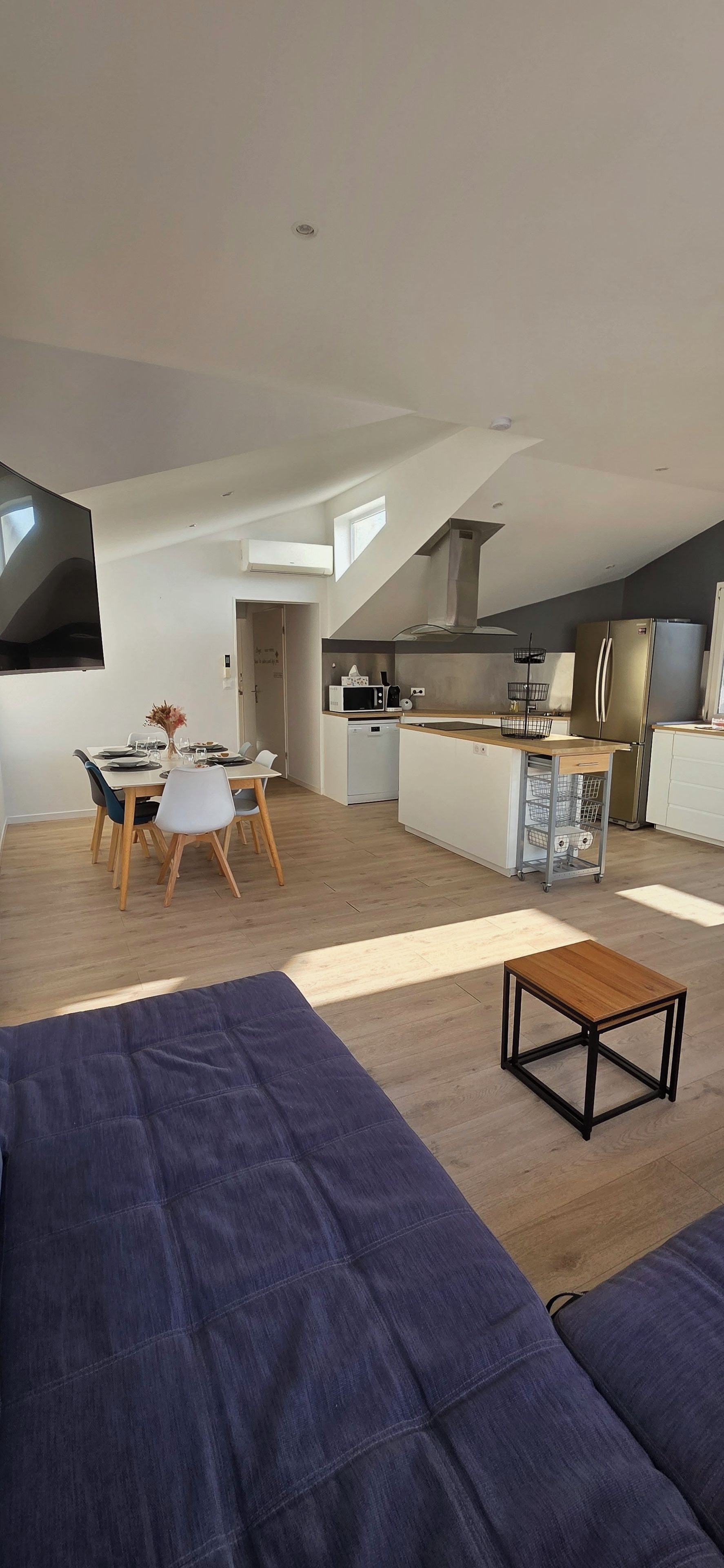 A modern living room with a sofa, a dining table with chairs, a kitchen area with appliances, and a large TV.