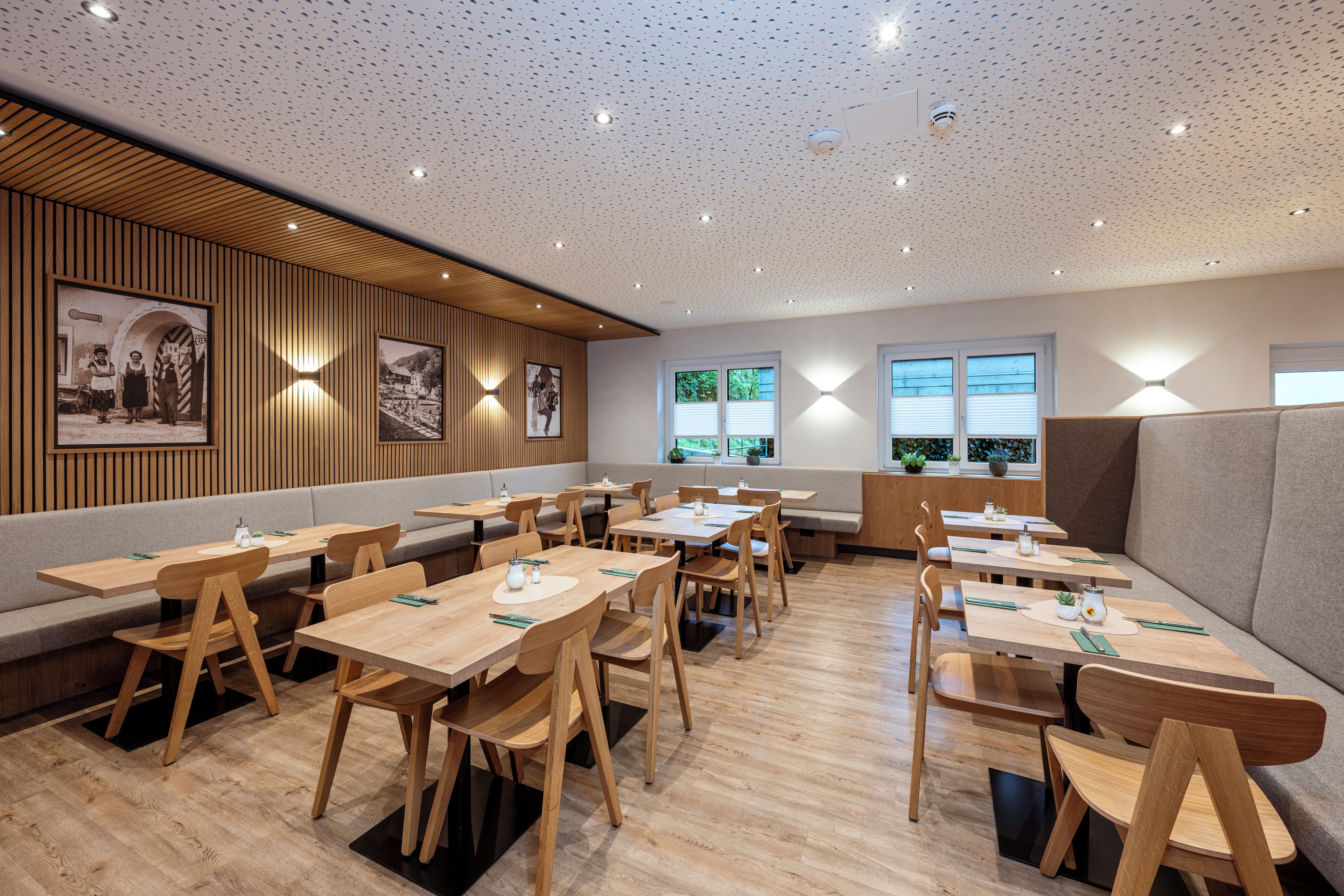 Interior de restaurante moderno com mesas e cadeiras de madeira, iluminação embutida no teto e quadros fixados na parede.