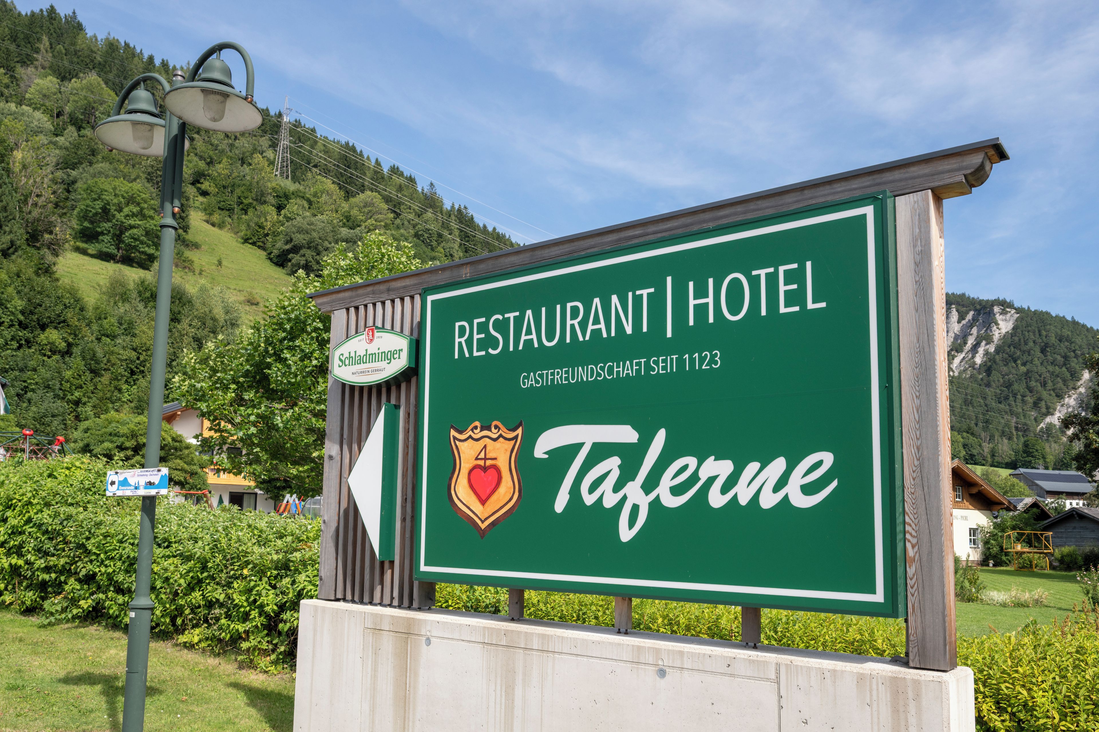 Schild für ein Restaurant und ein Hotel namens 'Taferne' mit Bergpanorama.