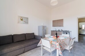 A modern living room with a grey sofa, a dining table set for four, and a kitchen area with a refrigerator and stove. - Residence La Baia - Bilo PT veranda by BarbarHouse (Casalabate)