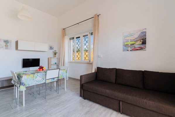 Ein Wohnzimmer mit einer Couch, einem Esstisch mit Stühlen, einem Fernseher und einem Fenster mit Vorhängen.