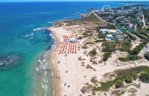 Vista aérea de una zona costera con playa, una fila de sombrillas y un conjunto de edificios.