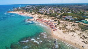 Luftaufnahme einer Küstenstadt mit einem Strand, der mit orangefarbenen Sonnenschirmen gesäumt ist, einem klaren, blauen Meer und Wohnhäusern im Hintergrund.