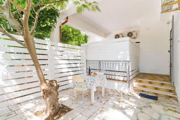 Ein überdachter Patio-Bereich mit weißem Zaun, einem Tisch mit Stühlen und einer Treppe, die zu einer erhöhten Ebene führt.