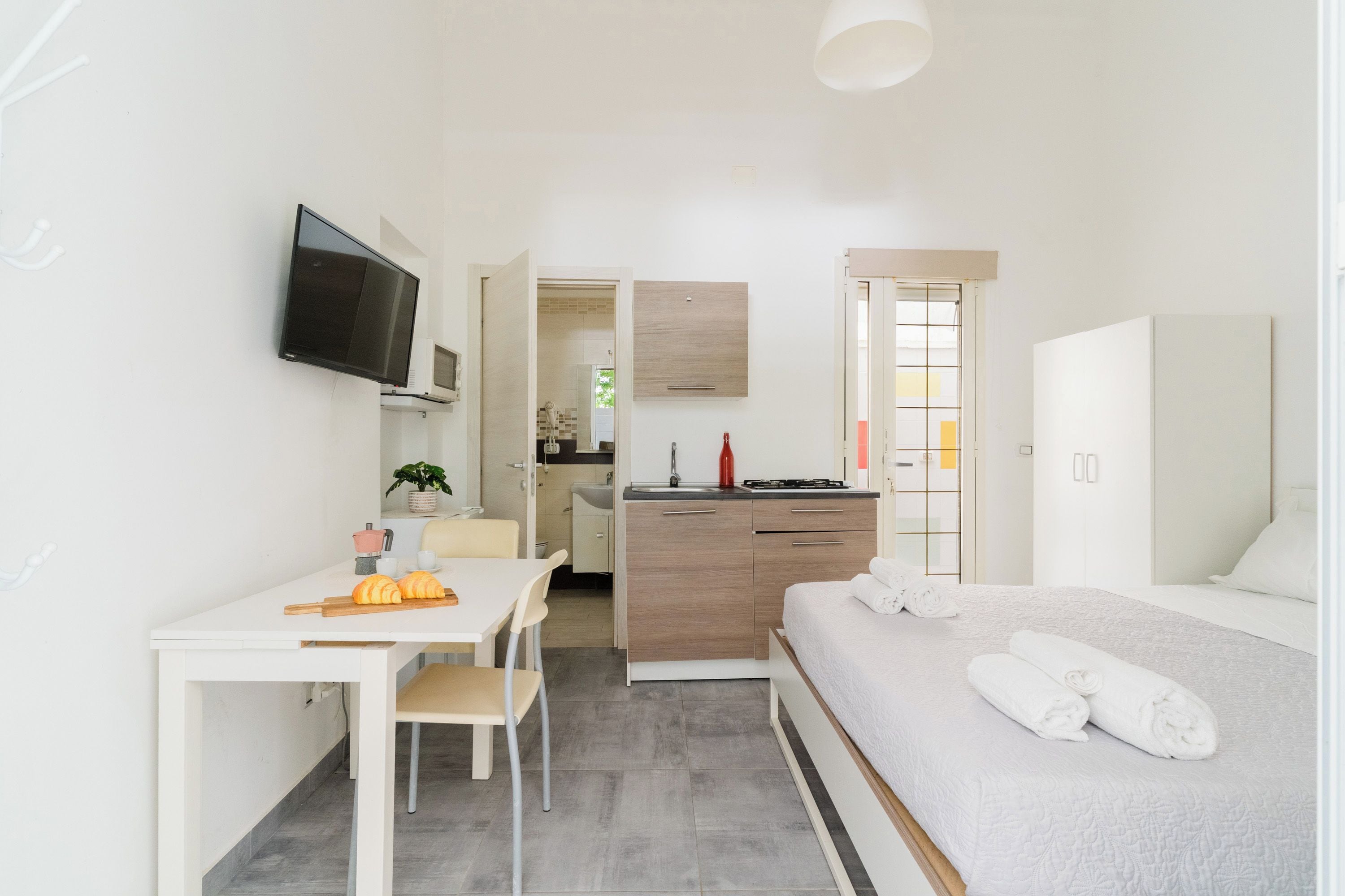 Habitación moderna y bien iluminada con cama, escritorio con silla, kitchenette y televisor montado en la pared.