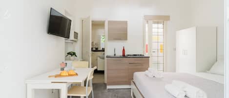 Habitación moderna y bien iluminada con cama, escritorio con silla, kitchenette y televisor montado en la pared.