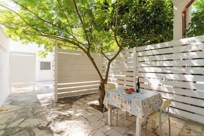 Un patio con mesa lista para comer, cerca blanca y un árbol que da sombra.