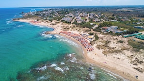An aerial view of a coastal town with a beach lined with orange umbrellas, a clear blue sea, and residential buildings in the background. - Residence La Baia-Trilo PP Standard by BarbarHouse (Casalabate)