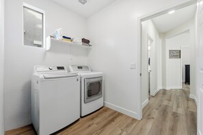 Una sala de bugaderia amb dues màquines blanques, una estant amb tovalloles pliegades i un passadís que condueix a una altra habitació.