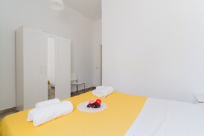 Ein Hotelzimmer mit einem Bett, einer gelben Tagesdecke, weißen Handtüchern, einem Teller mit Obst und einem Spiegel.