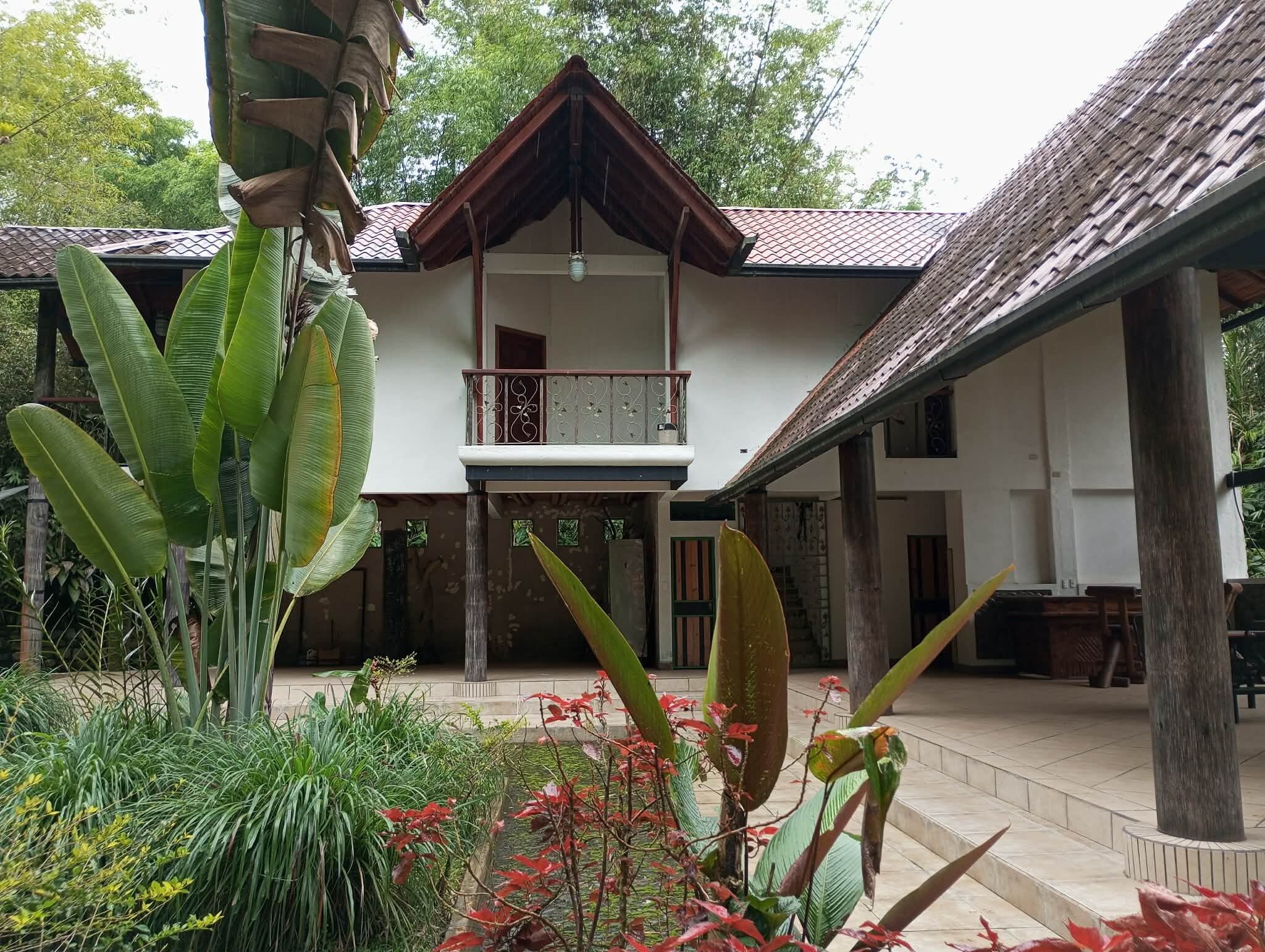 A two-story building with a balcony, surrounded by tropical plants and a tiled roof.