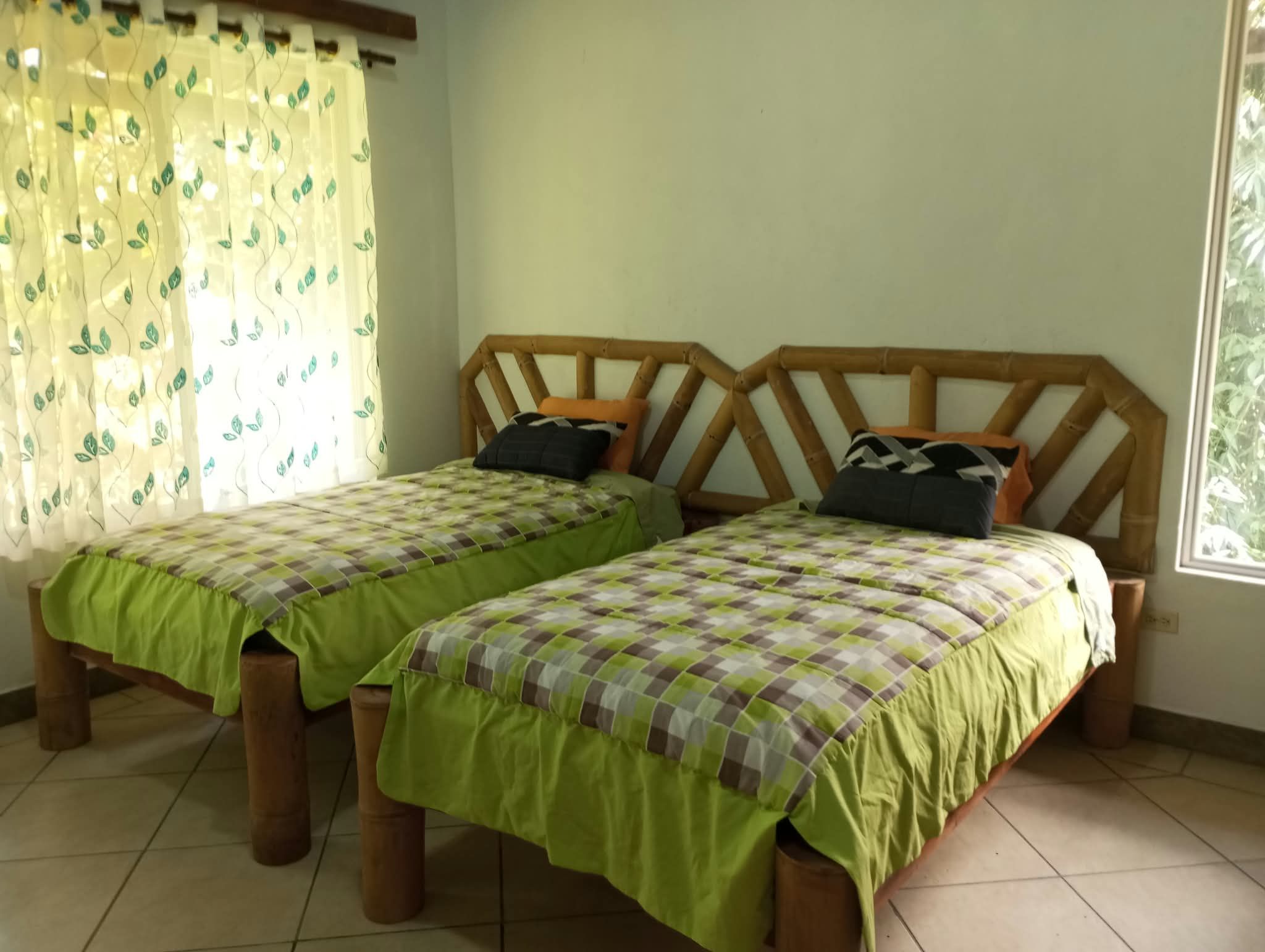 A room with two wooden beds with checkered bedding, a bamboo headboard, and a window with a curtain.