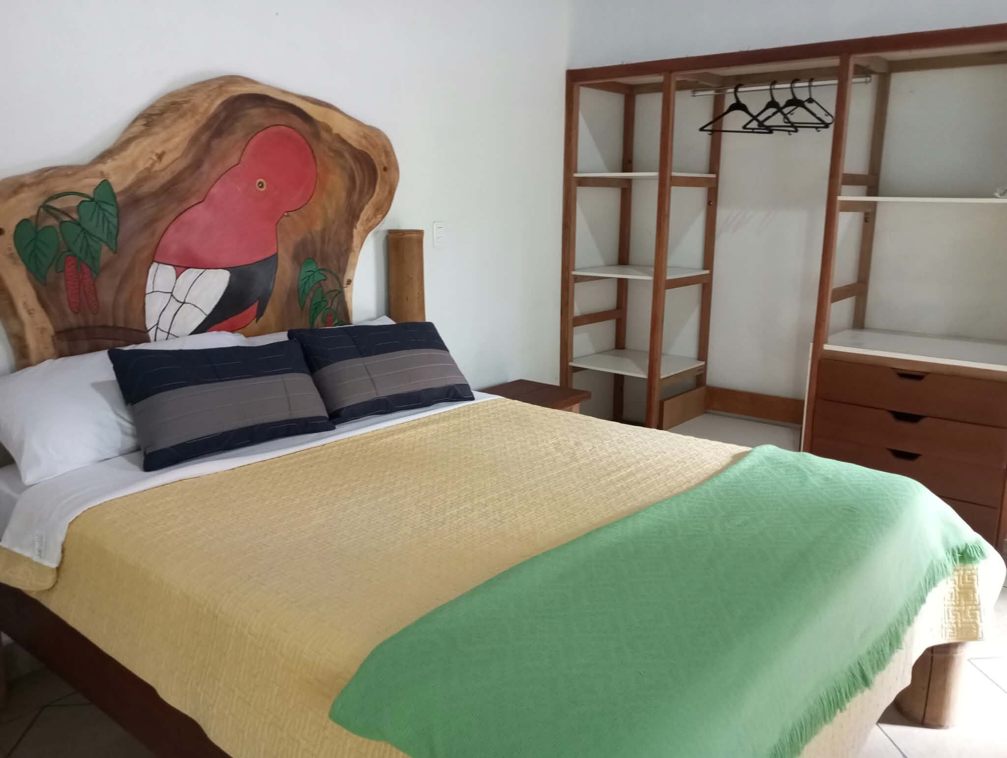 A bedroom with a bed, pillows, a green throw blanket, a wooden headboard with a bird design, a wardrobe with hanging space, and a nightstand.
