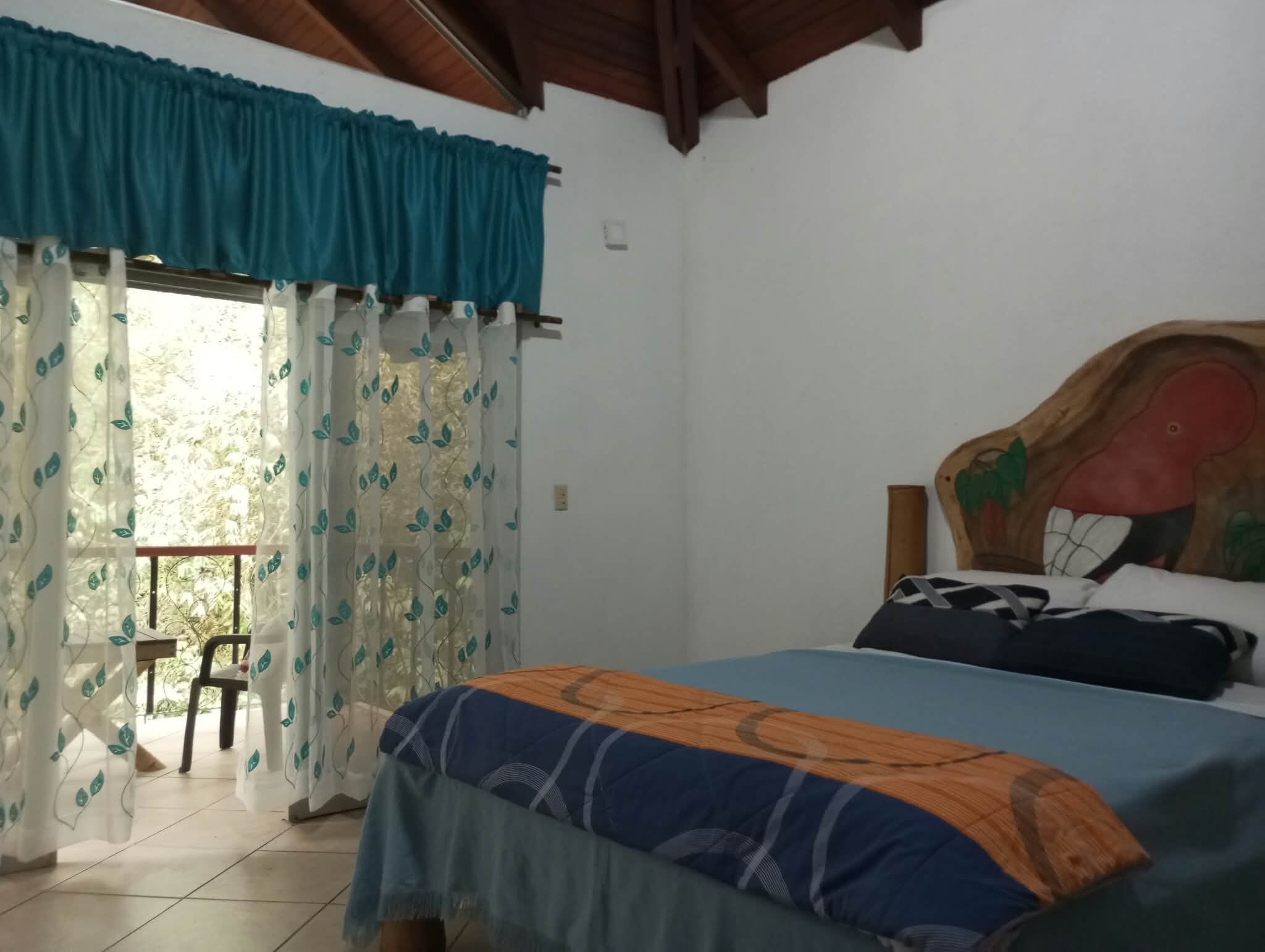 Un dormitorio con cama, una silla, un ventanal con cortinas y techo de madera.