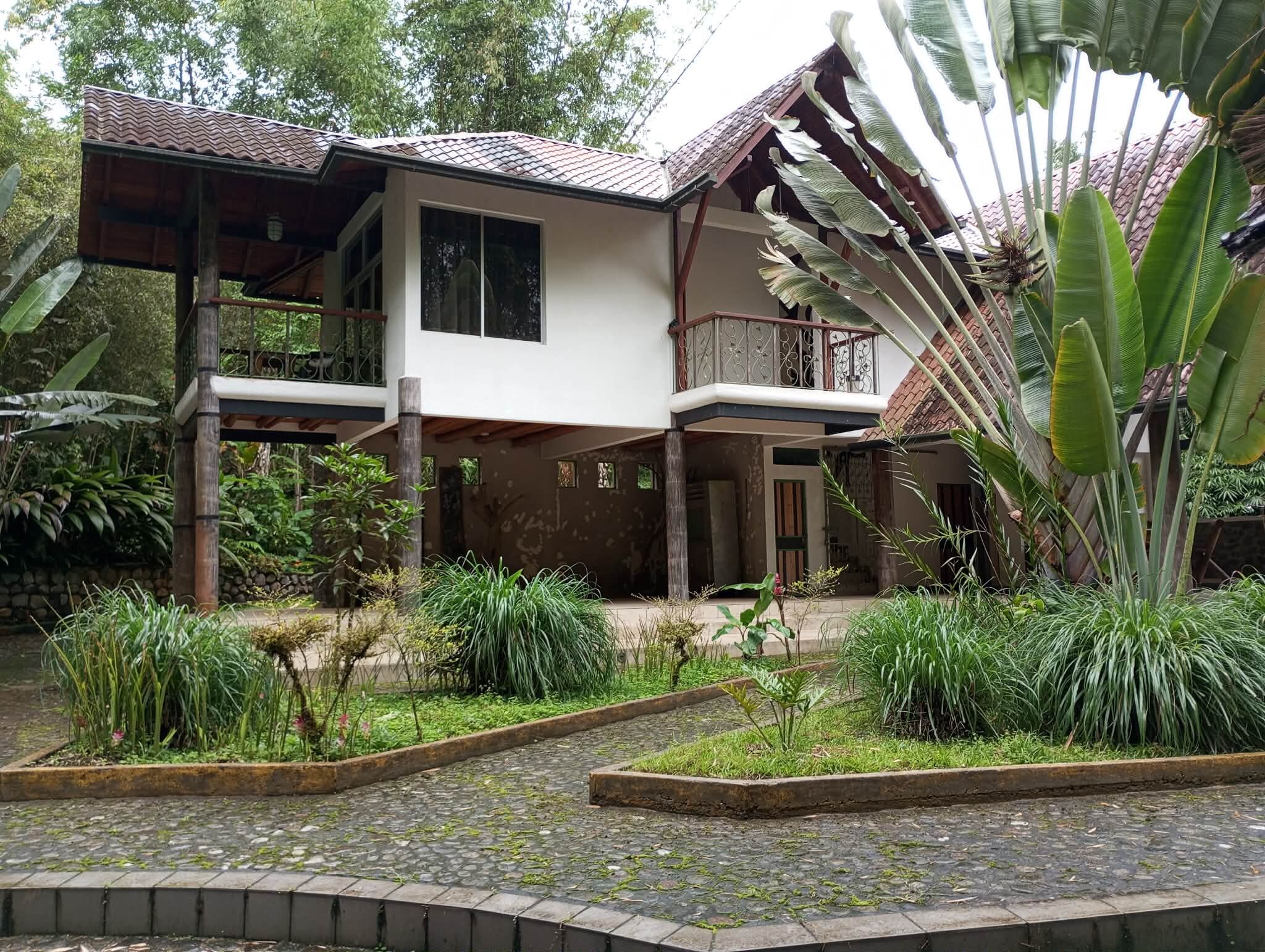 A two-story house with a balcony, surrounded by lush greenery and a cobblestone pathway.