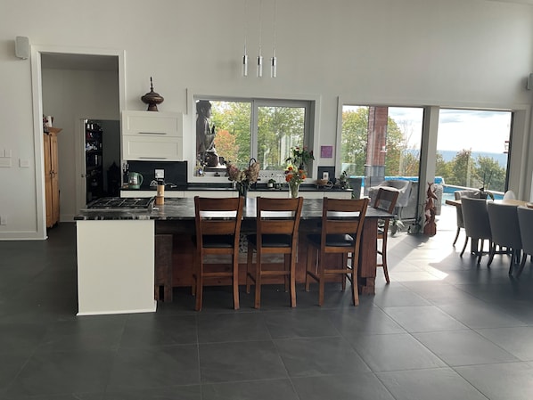 A modern kitchen with a central island, dining area with chairs, and large windows offering a view of the outdoors.