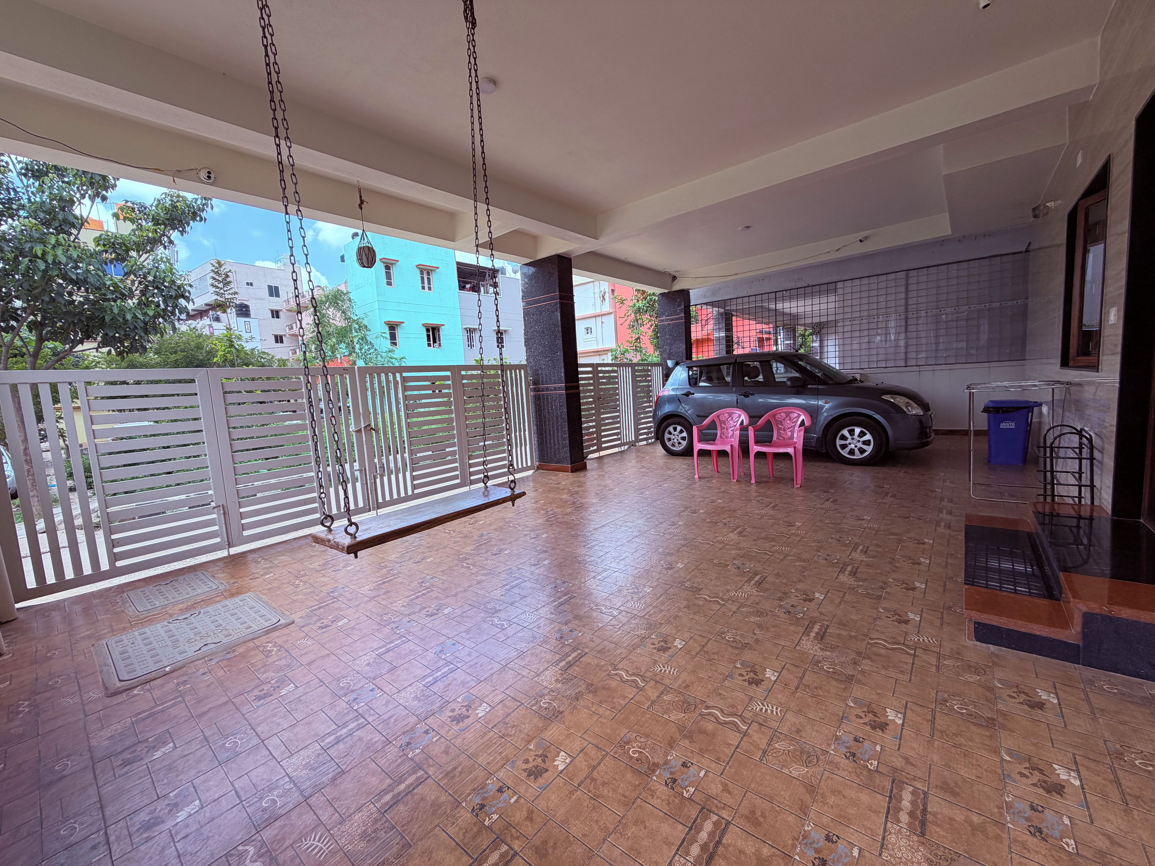 A covered outdoor area with a swing, two pink chairs, a parked car, and a blue trash bin.
