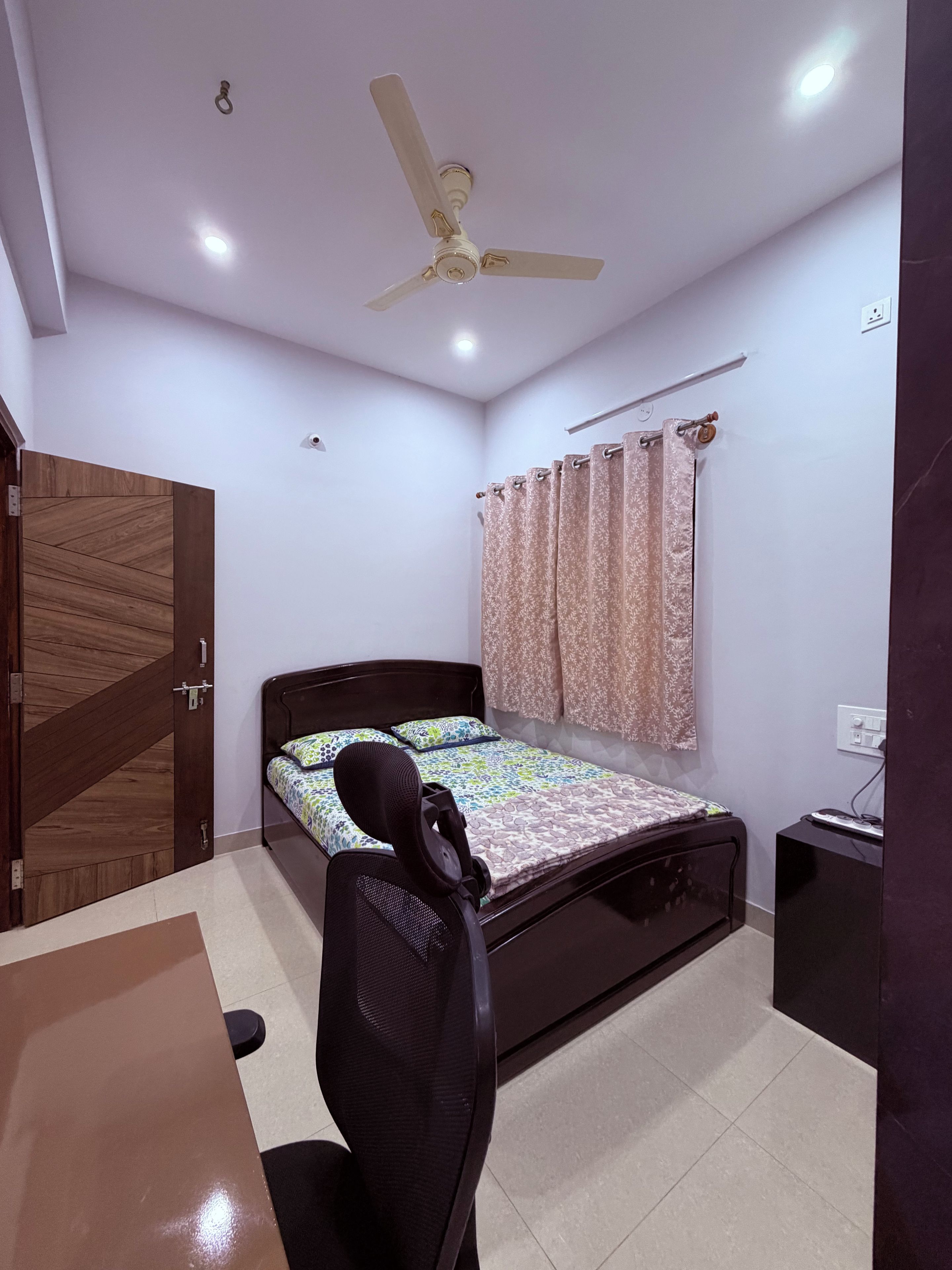 A bedroom with a bed, a desk, a chair, a ceiling fan, and a window with curtains.