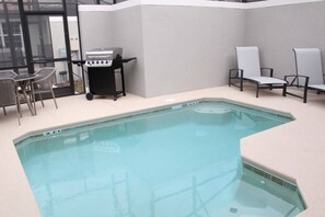 Une piscine intérieure dotée d’un barbecue, de chaises longues et d’une porte vitrée donnant sur l’extérieur.