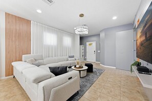 A modern living room with a sectional sofa, a chandelier, and a flat-screen TV mounted on the wall. - 5BR Disney-area Home Pool, Theater, Themed (Kissimmee)