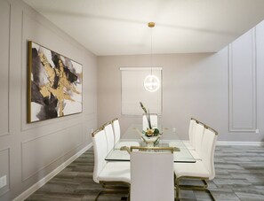 A dining area with a glass table, white chairs, a modern light fixture, and a large abstract painting on the wall. - Themed 5BR Near Disney Pool, Game Room (Kissimmee)