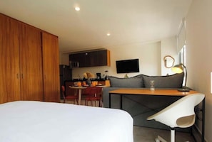 Quarto de hotel compacto com cama, cozinha compacta, espaço para refeições e área de estar com mesa e cadeiras.