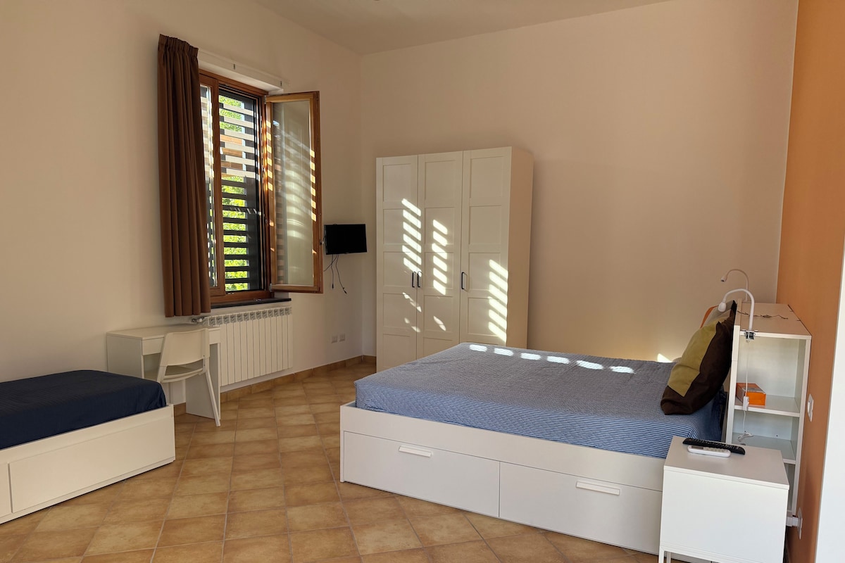 A bedroom with a bed, a desk, a chair, a wardrobe, and a bedside table with a lamp.